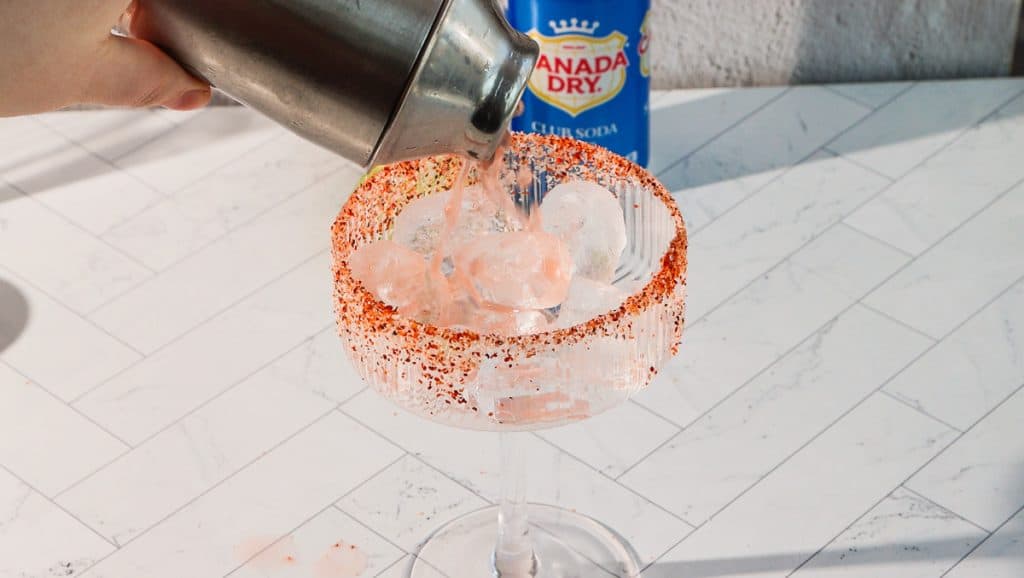 A pink spicy margarita mocktail is poured into a glass with ice and a chili powder rim, with a Canada Dry club soda can nearby.