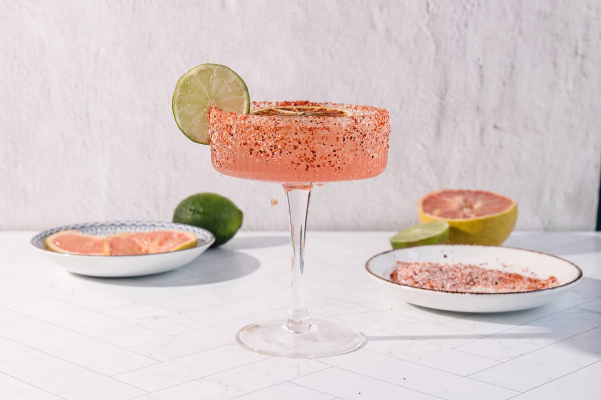 A pink Spicy Grapefruit Margarita Mocktail with a chili rim, garnished with a lime slice, sits on a white surface. This refreshing non-alcoholic drink is perfect for any occasion.