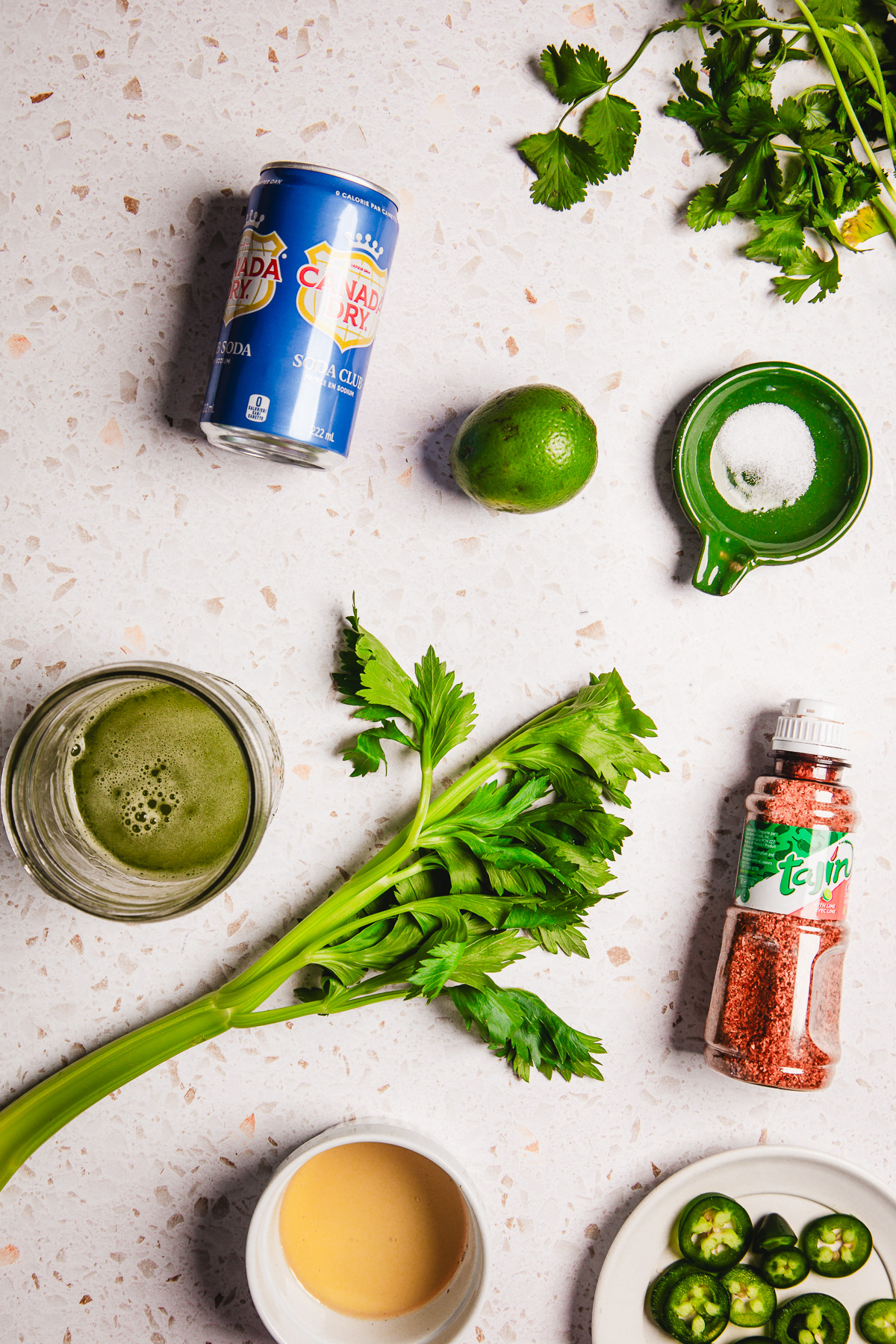 Celery, lime, club soda, cilantro, jalapeño, salt, chili powder, green juice, and a bowl of light-colored sauce—perfect ingredients for a refreshing Spicy Margarita Mocktail or another zesty non-alcoholic cocktail.