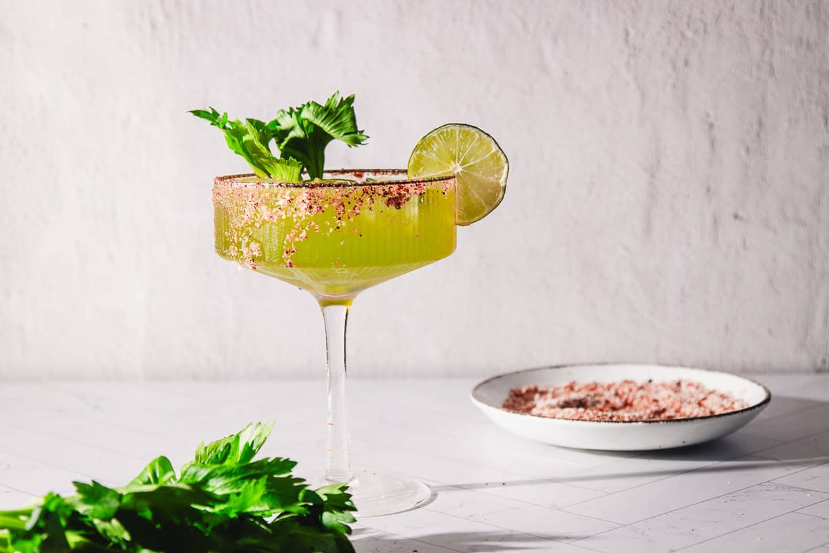 A Margarita Mocktail with a salted rim, lime slice, and celery garnish sits ready to enjoy, with salt on a plate and fresh celery nearby.