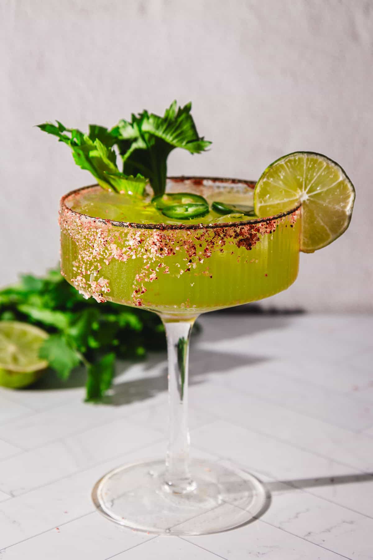 A green Spicy Margarita Mocktail in a glass rimmed with spice, garnished with celery, jalapeño slices, and a lime wheel.