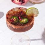 A spicy pomegranate mocktail garnished with lime, jalapeño slices, and pomegranate seeds, served in a glass with a spiced rim.