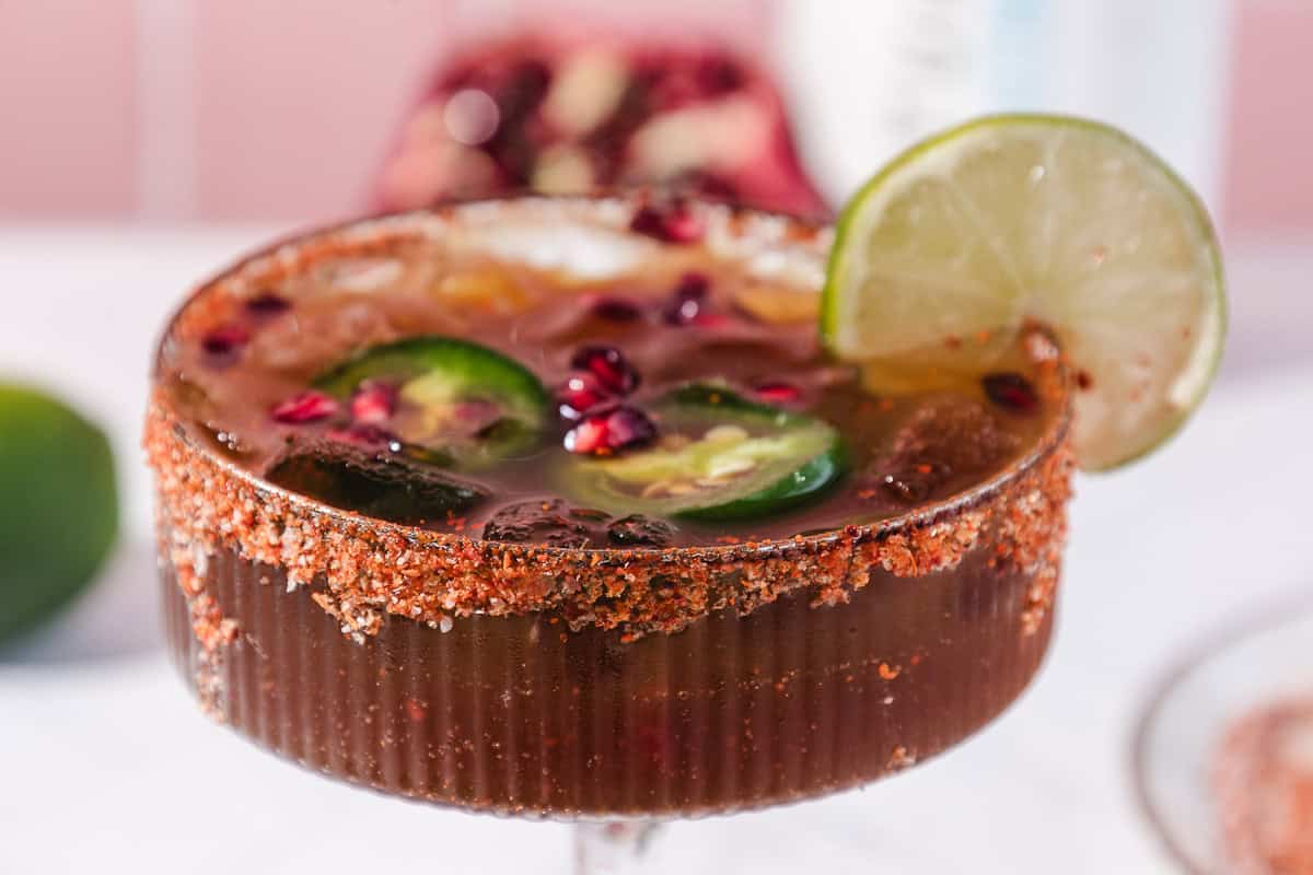 A spicy pomegranate mocktail served in a chili-rimmed glass, garnished with lime slice, jalape&ntilde;o, pomegranate seeds, and ice cubes.