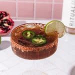 A spicy pomegranate mocktail with lime and jalape&ntilde;o slices in a glass with a chili rim, served beside fresh lime and pomegranate.