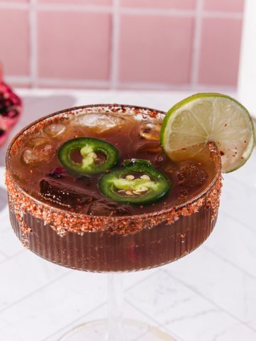 A spicy pomegranate mocktail with lime and jalape&ntilde;o slices in a glass with a chili rim, served beside fresh lime and pomegranate.