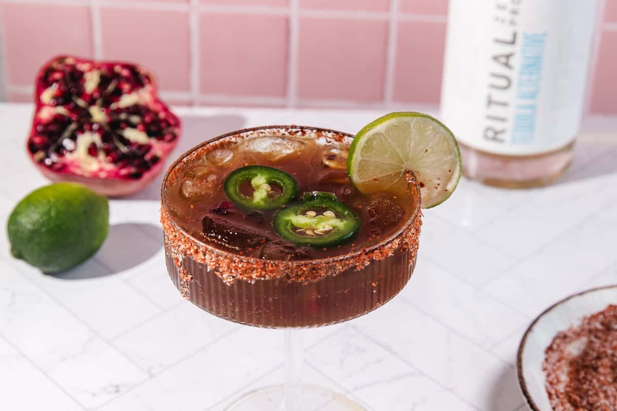 A spicy pomegranate mocktail with lime and jalape&ntilde;o slices in a glass with a chili rim, served beside fresh lime and pomegranate.