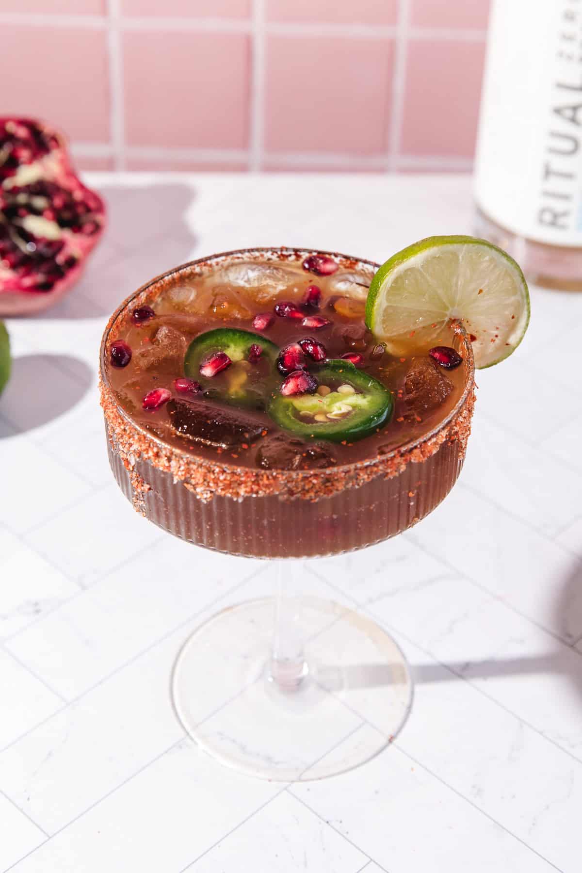 A spicy pomegranate mocktail with ice, pomegranate seeds, jalape&ntilde;o slices, a lime wheel, and a chili-rimmed glass.
