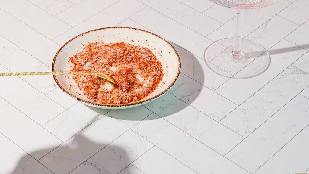 A bowl of red and white seasoning mix with a spoon sits next to a wine glass filled with a spicy pomegranate mocktail on a white tiled surface.