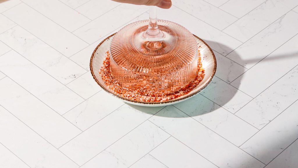 A glass, ready for a spicy pomegranate mocktail, sits upside down in a bowl of chili powder and salt on a white tile surface.
