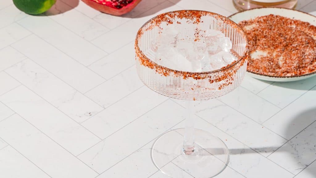 A cocktail glass with ice and a chili powder rim sits on a white countertop near a plate of chili powder, ready for a spicy pomegranate mocktail.