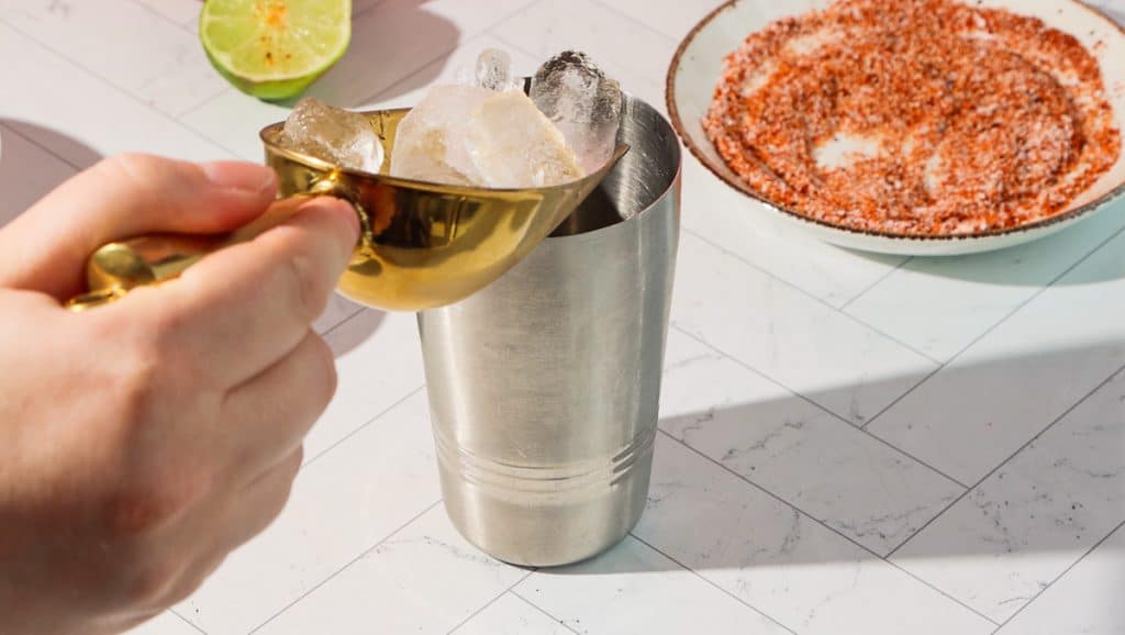 A hand scoops ice cubes into a metal cocktail shaker, ready to craft a spicy pomegranate mocktail, with a sliced lime and chili salt nearby.