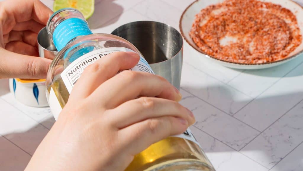 A person pours liquid from a bottle into a metal shaker near a plate with a spicy seasoning mix, preparing to craft a spicy pomegranate mocktail.