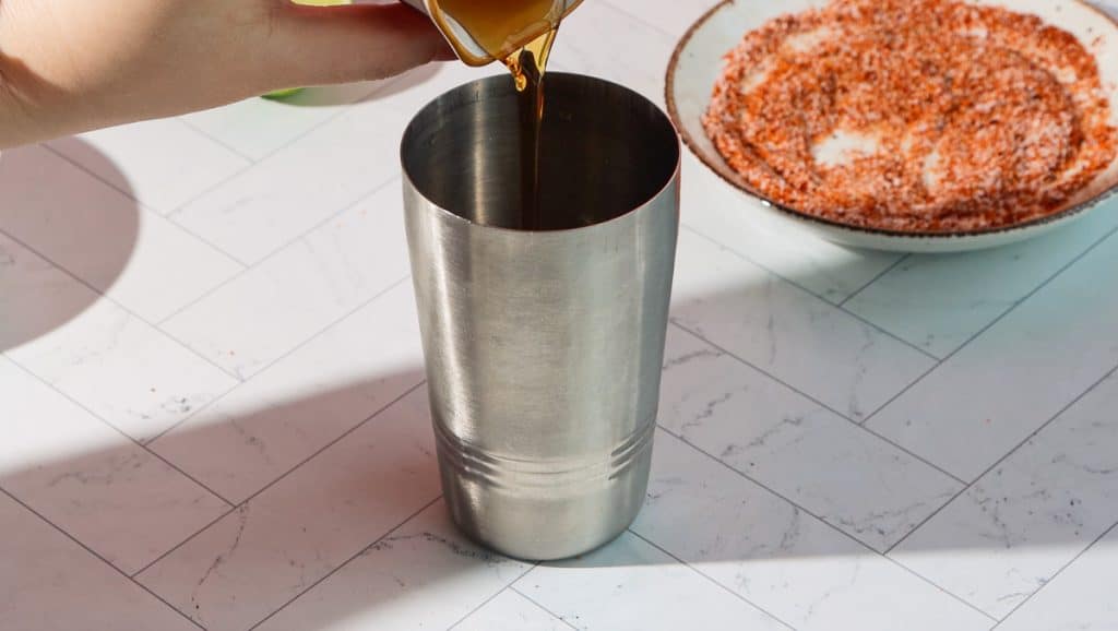 A hand pours liquid into a metal shaker for a spicy pomegranate mocktail, with a plate of spice mix in the background on a tiled surface.