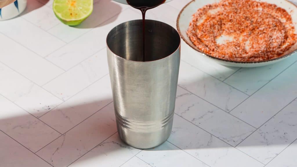 A metal cocktail shaker being filled with liquid; lime and chili powder on a plate nearby, perfect for crafting a spicy pomegranate mocktail.