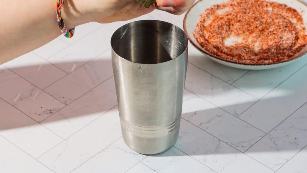 A hand holds a lime over a metal shaker while preparing a spicy pomegranate mocktail, with a plate of spiced salt in the background.