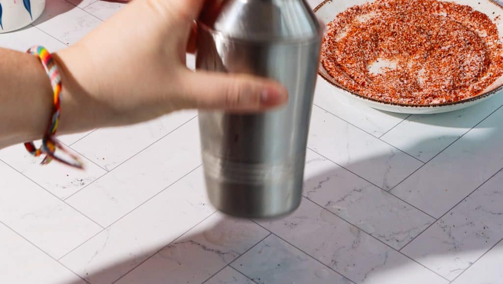 Hand holding a cocktail shaker near a plate with red seasoning, ready to craft a spicy pomegranate mocktail on a white tiled surface.