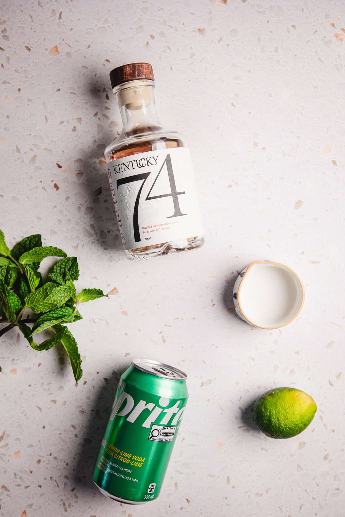 A bottle of Kentucky 74, a can of Sprite, a lime, mint leaves, and a small cup on a light surface&mdash;perfect for crafting your favorite virgin mint julep recipe.