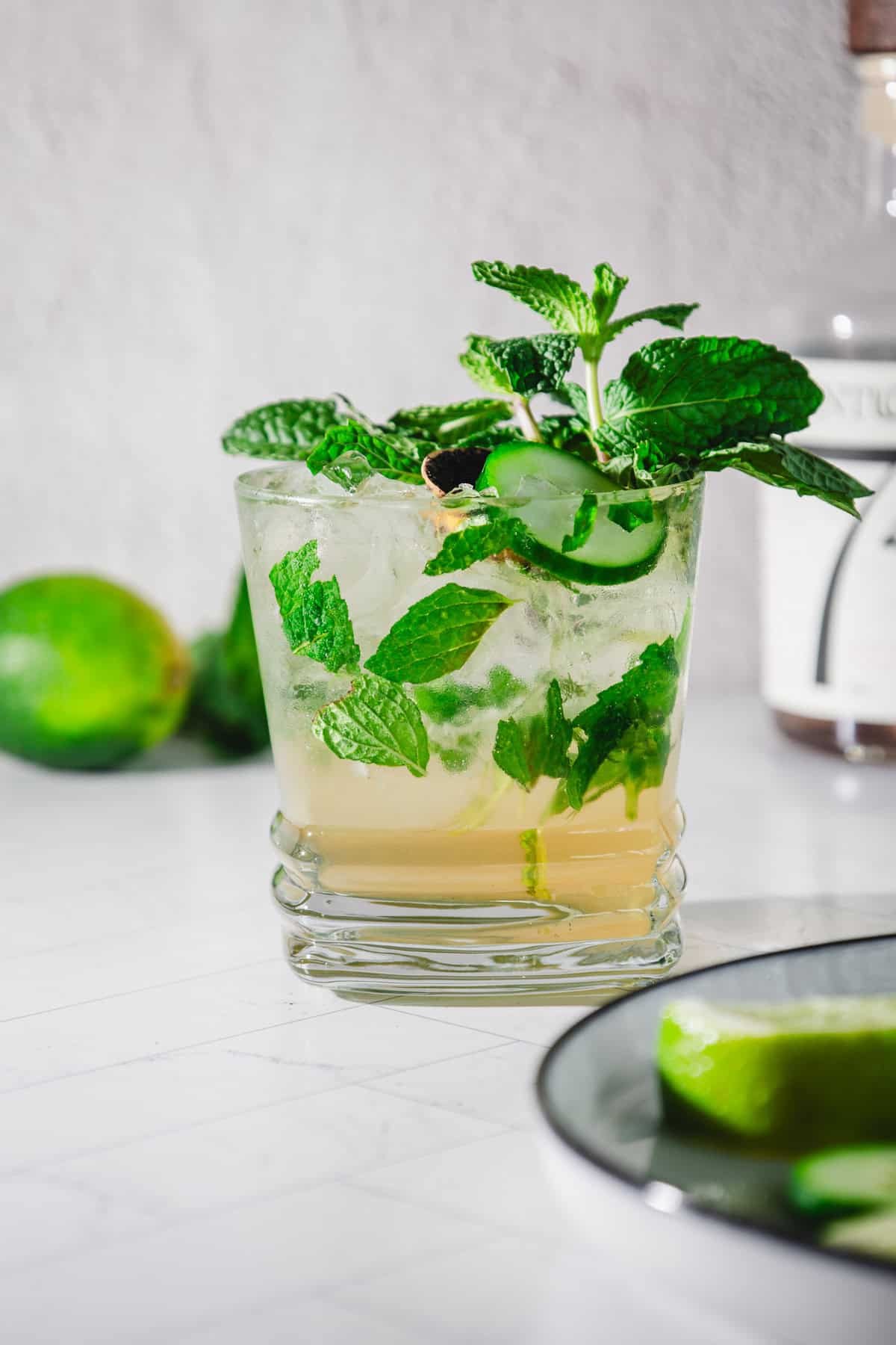 A glass of mojito with mint leaves, lime slices, and ice sits in front of a whole lime and bottle&mdash;perfect inspiration for a refreshing virgin mint julep recipe.