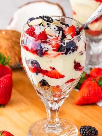 Four parfait glasses with layered berries and cream, surrounded by fresh strawberries on a wooden board.