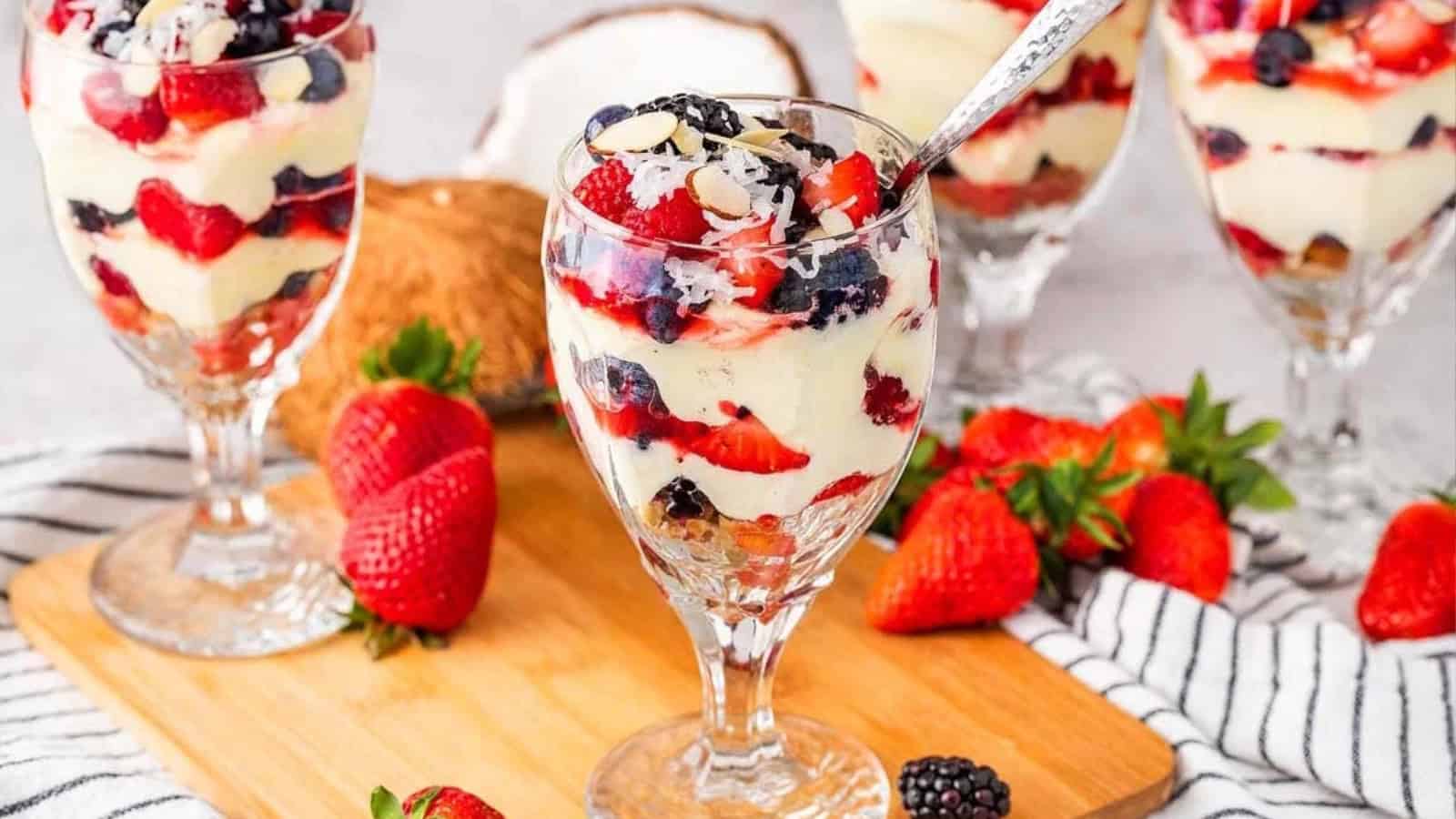 Four parfait glasses with layered berries and cream, surrounded by fresh strawberries on a wooden board.