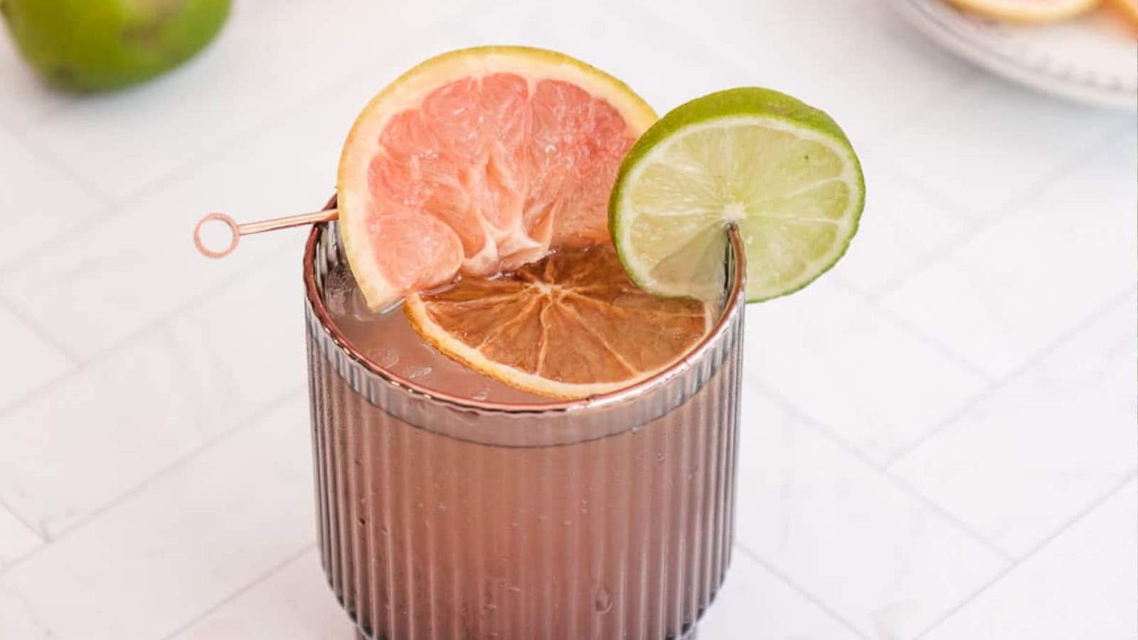 A cocktail garnished with grapefruit, lime, and dried orange slices in a ribbed glass on a white surface.