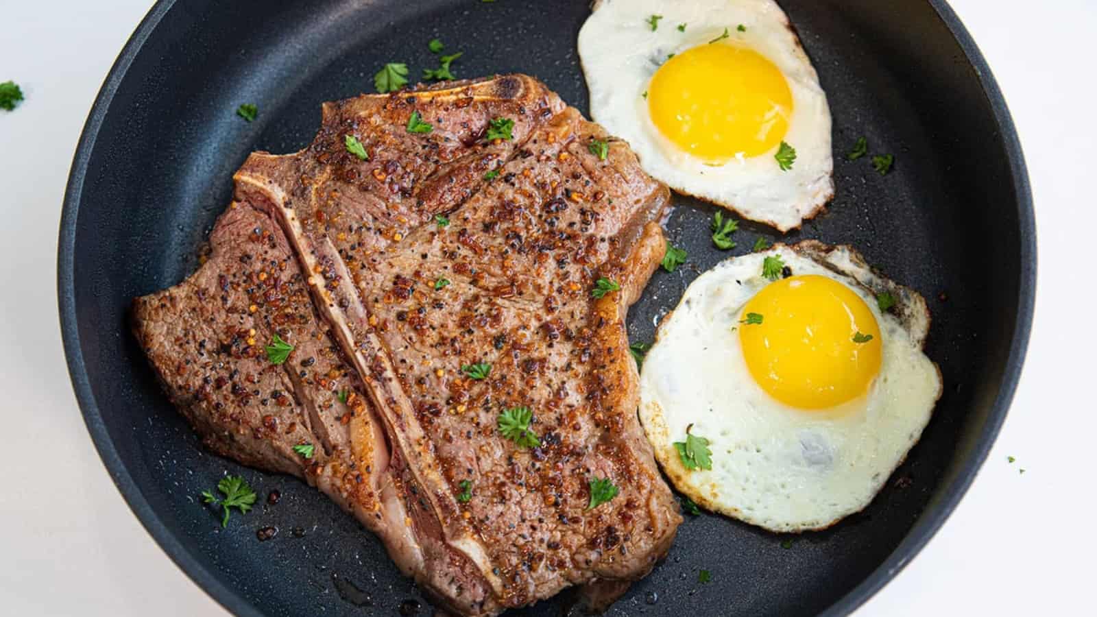A cooked T-bone steak and two sunny-side-up eggs in a black frying pan, garnished with parsley.