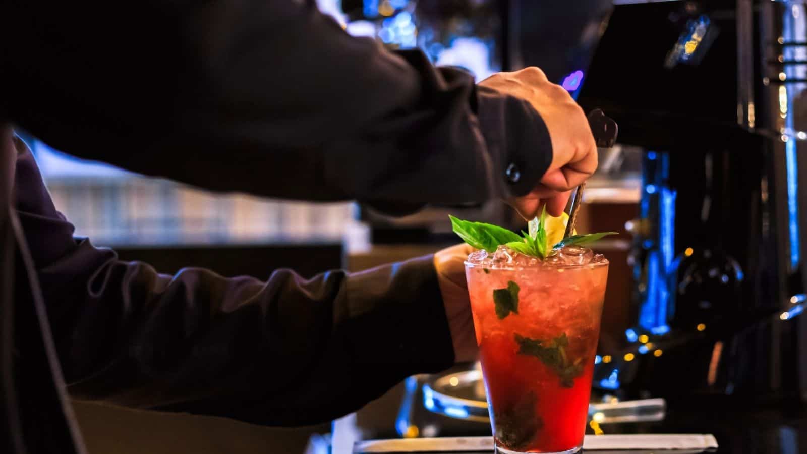 Bartender stirring a vibrant red mocktail with mint leaves and ice at a dimly lit bar counter, showcasing creative non-alcoholic drinks.