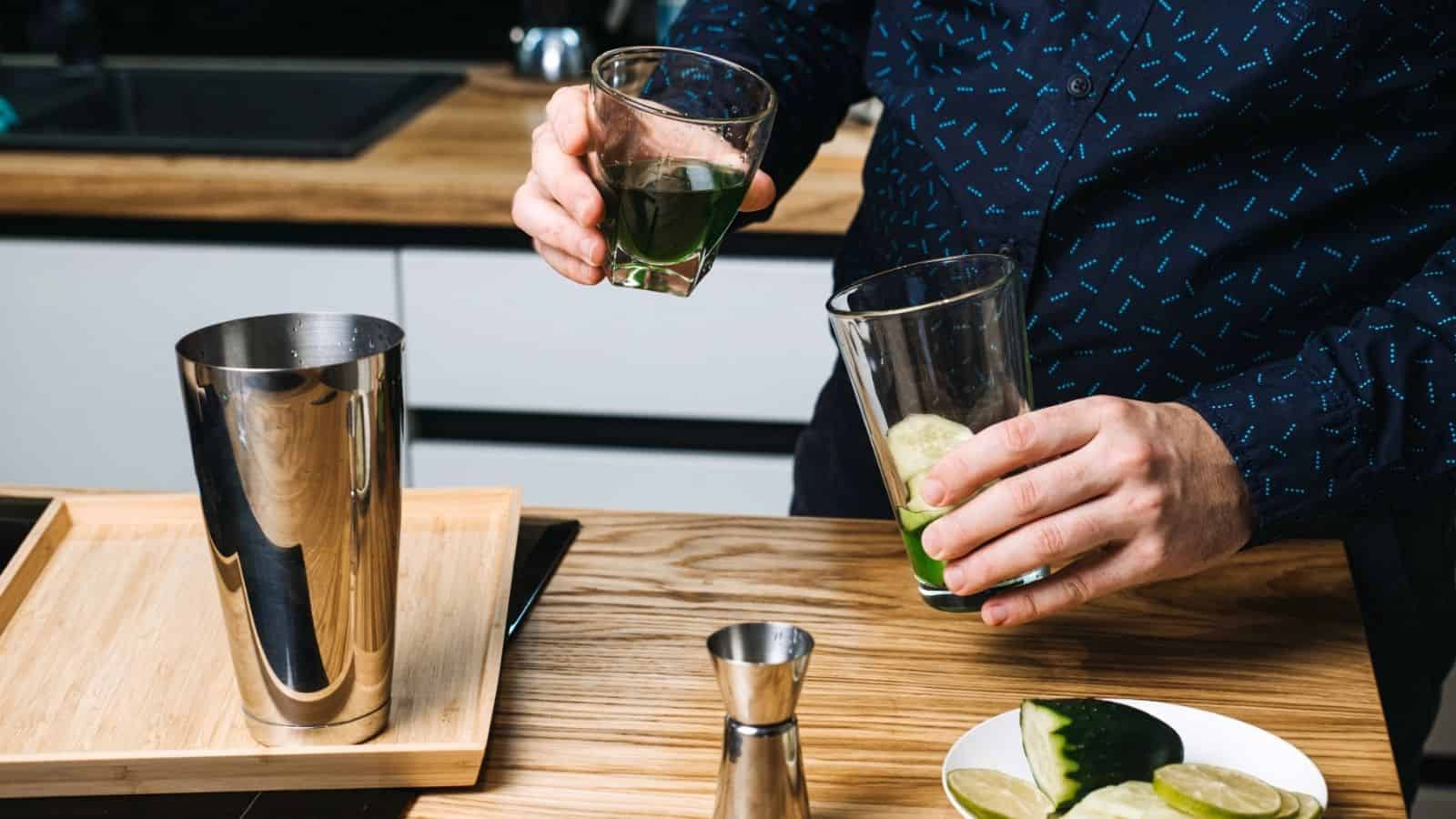 Person mixing a green mocktail in glasses with lime, near a shaker and lime slices on a kitchen counter&mdash;perfect for those learning how to make mocktails or enjoy non-alcoholic cocktails.