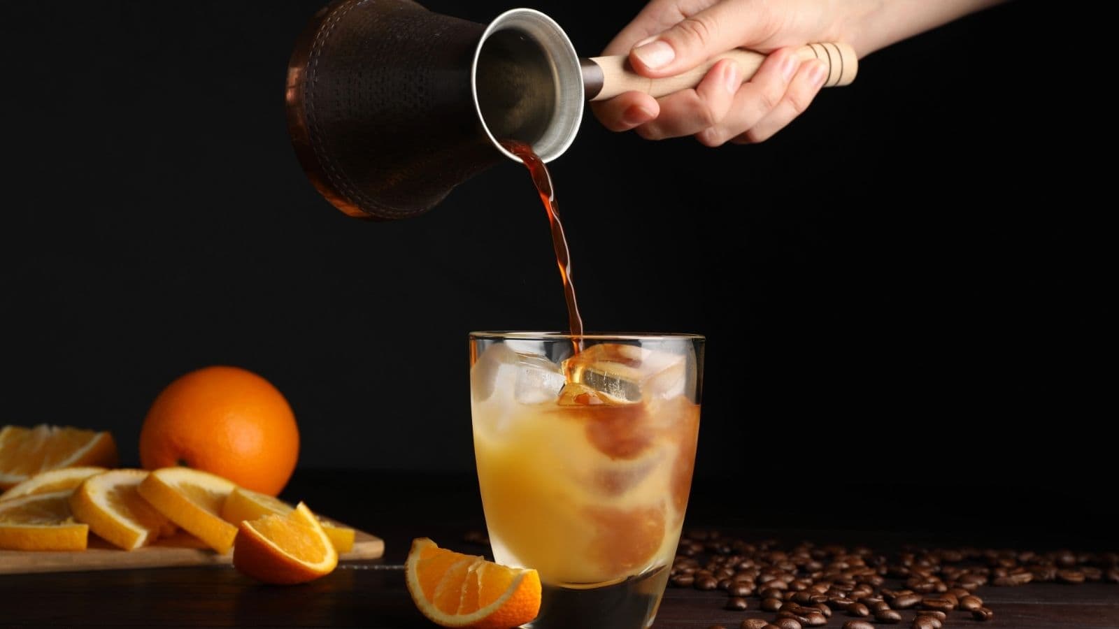 A hand pours coffee into an iced orange drink, creating one of the most refreshing mocktail recipes, with orange slices and coffee beans beside the glass.