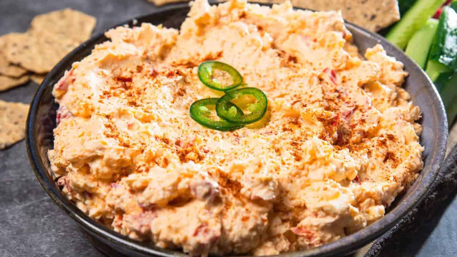 A bowl of creamy pimento cheese dip topped with sliced jalapeรฑos and a sprinkle of paprika.