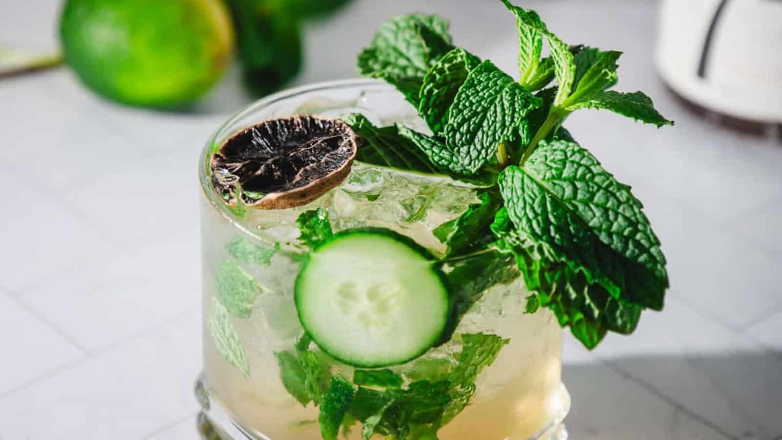 A cocktail garnished with mint, cucumber slice, and dried lime sits on a white surface.