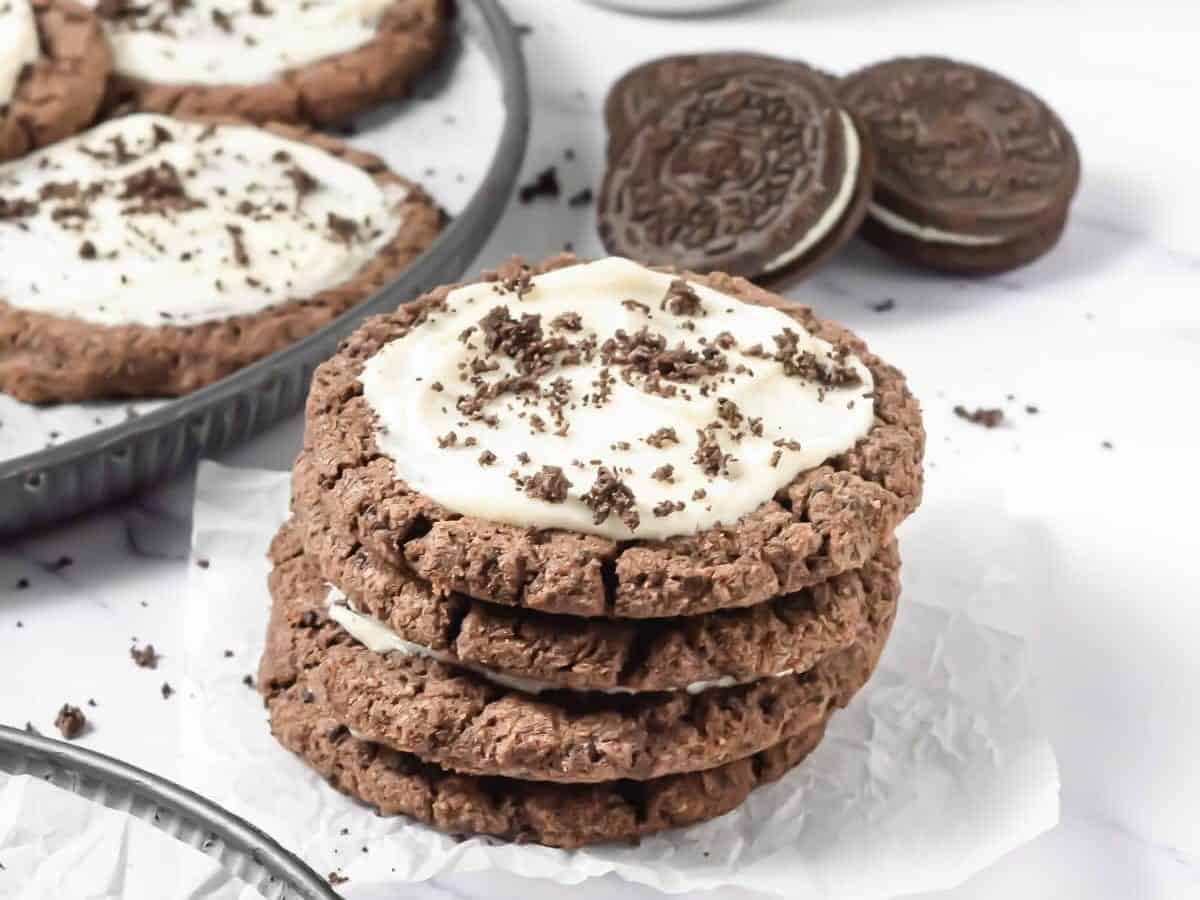 A stack of frosted Oreo cookies with white frosting and crumbs, next to classic chocolate sandwich cookiesโperfect for a Crumbl Copycat treat.