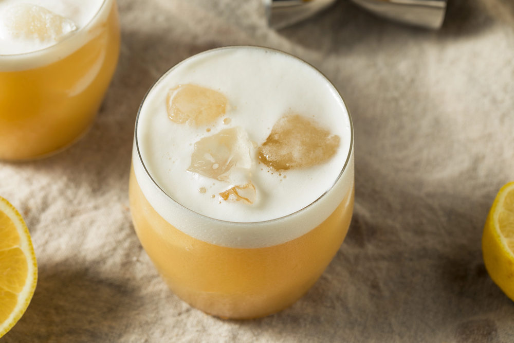 A glass of non-alcoholic tequila sour with ice and foam, set on a beige surface with lemon slices nearby.