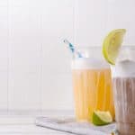 Three iced drinks with foam, lime wedges, and striped straws on a white surface with a light background.