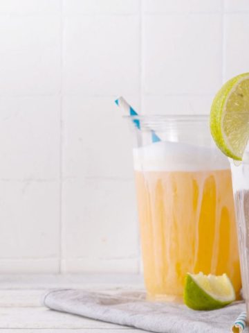 Three iced drinks with foam, lime wedges, and striped straws on a white surface with a light background.