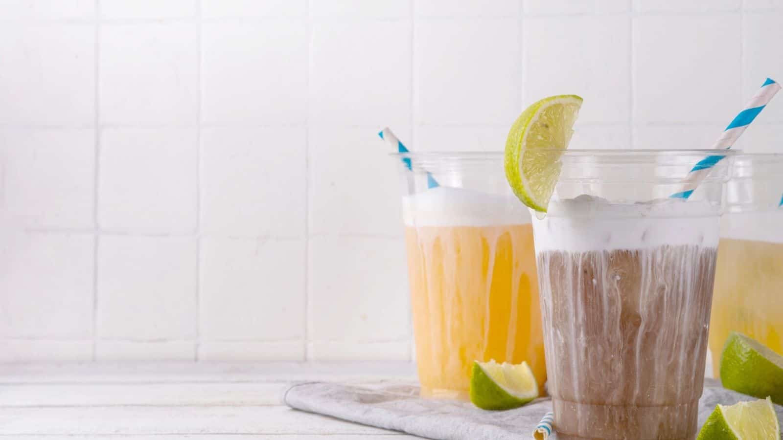Three iced drinks with foam, lime wedges, and striped straws on a white surface with a light background.