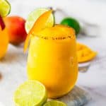 Bright frozen mango margarita mocktail in a glass, garnished with lime, mango slice, and chili rim, with fresh limes nearby.