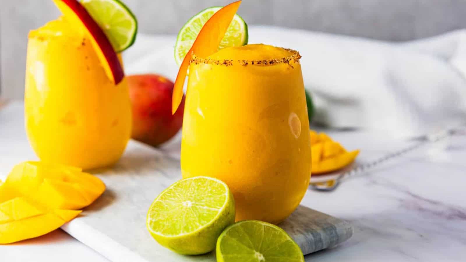 Two mango smoothies in glasses, garnished with lime and mango slices, on a white board with cut limes.