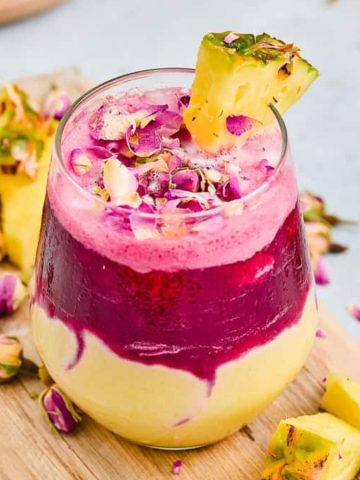 A glass of layered yellow and pink smoothie, topped with pineapple and rose petals on a wooden board.