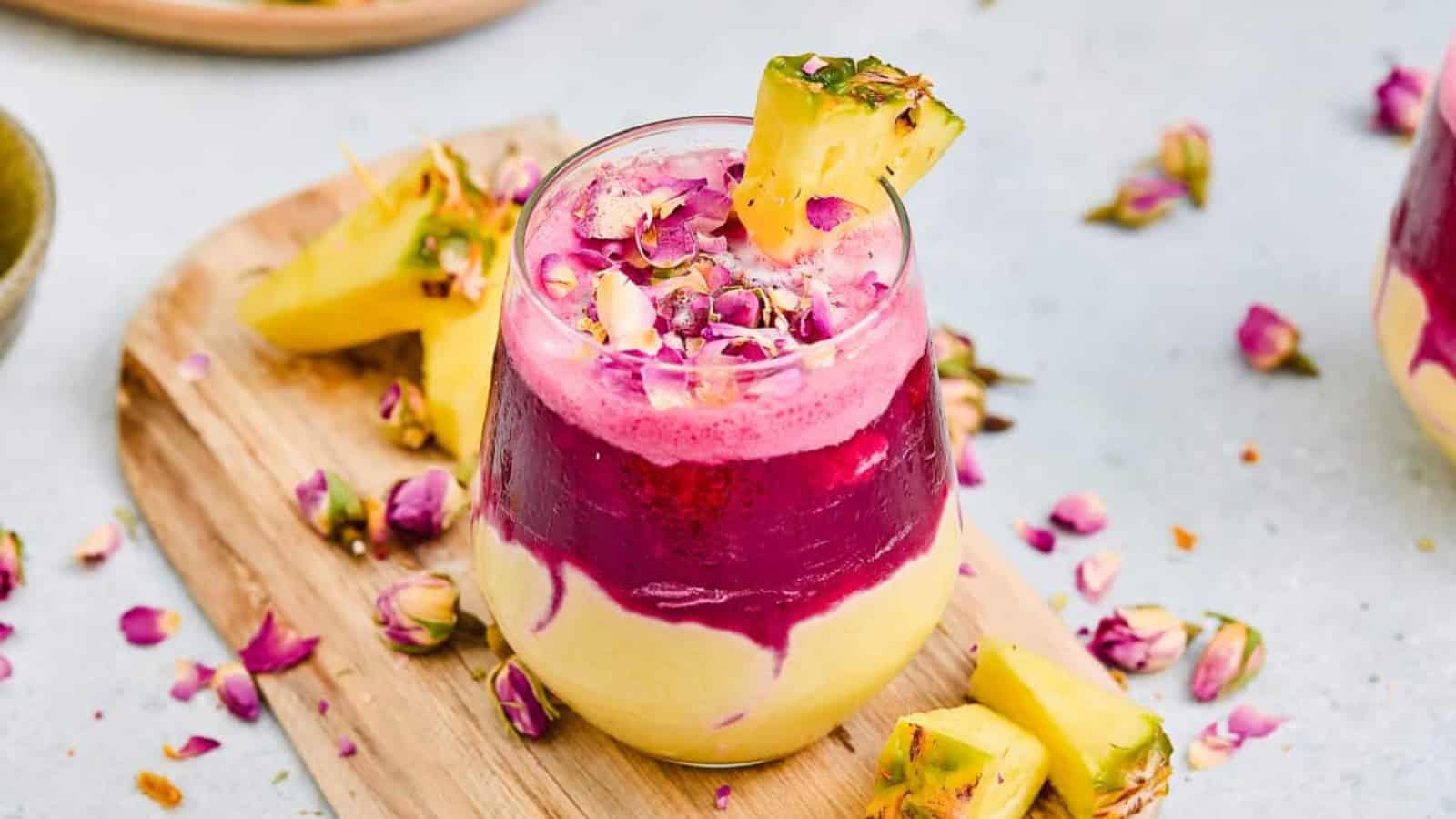 Layered smoothie with yellow and magenta swirls, topped with pineapple and dried flower petals on a wooden board.