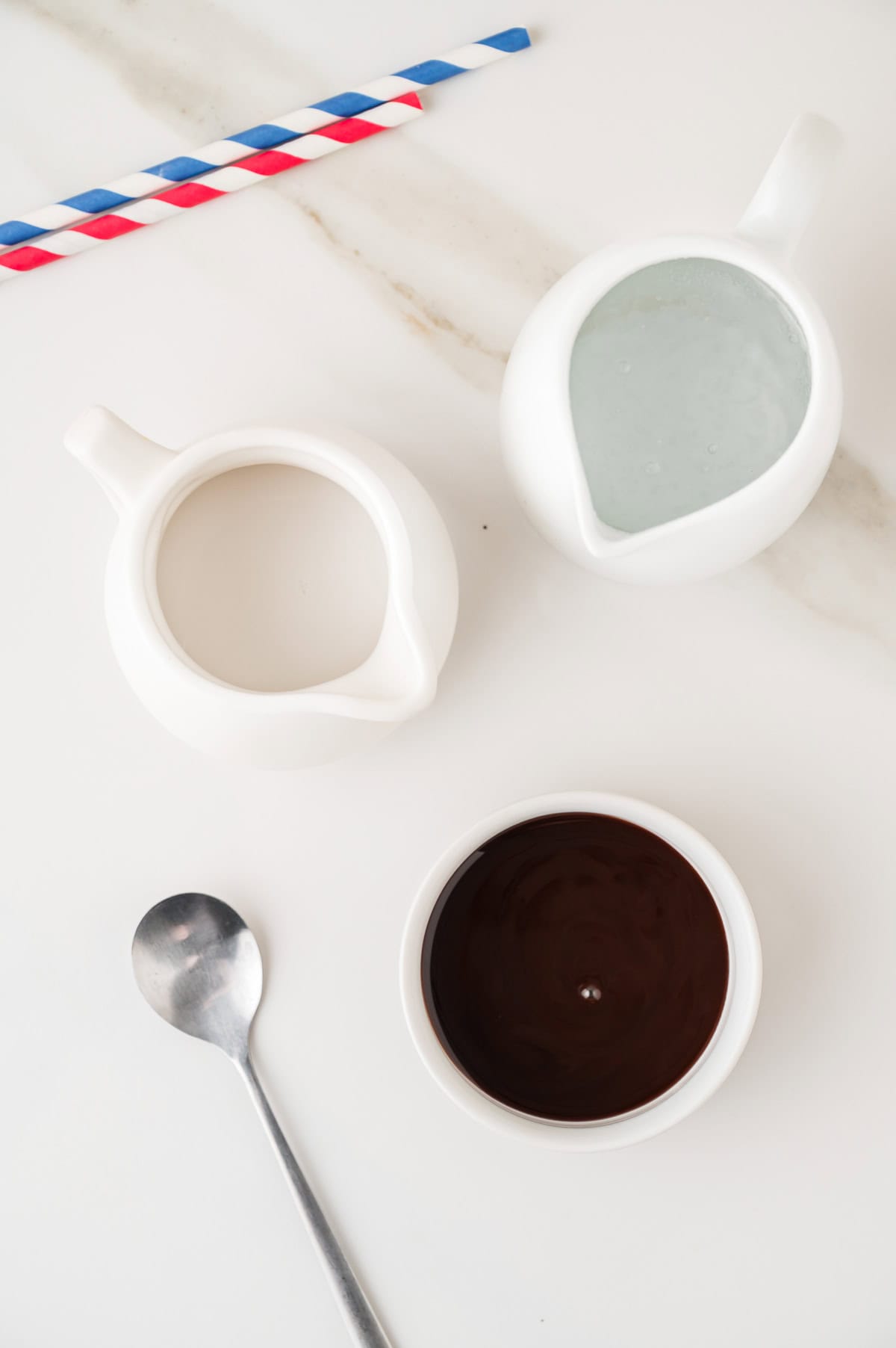 Two pitchers with milk, a bowl of chocolate sauce, a spoon, and two striped straws on a white surface evoke the classic preparation of an Egg Cream Soda.