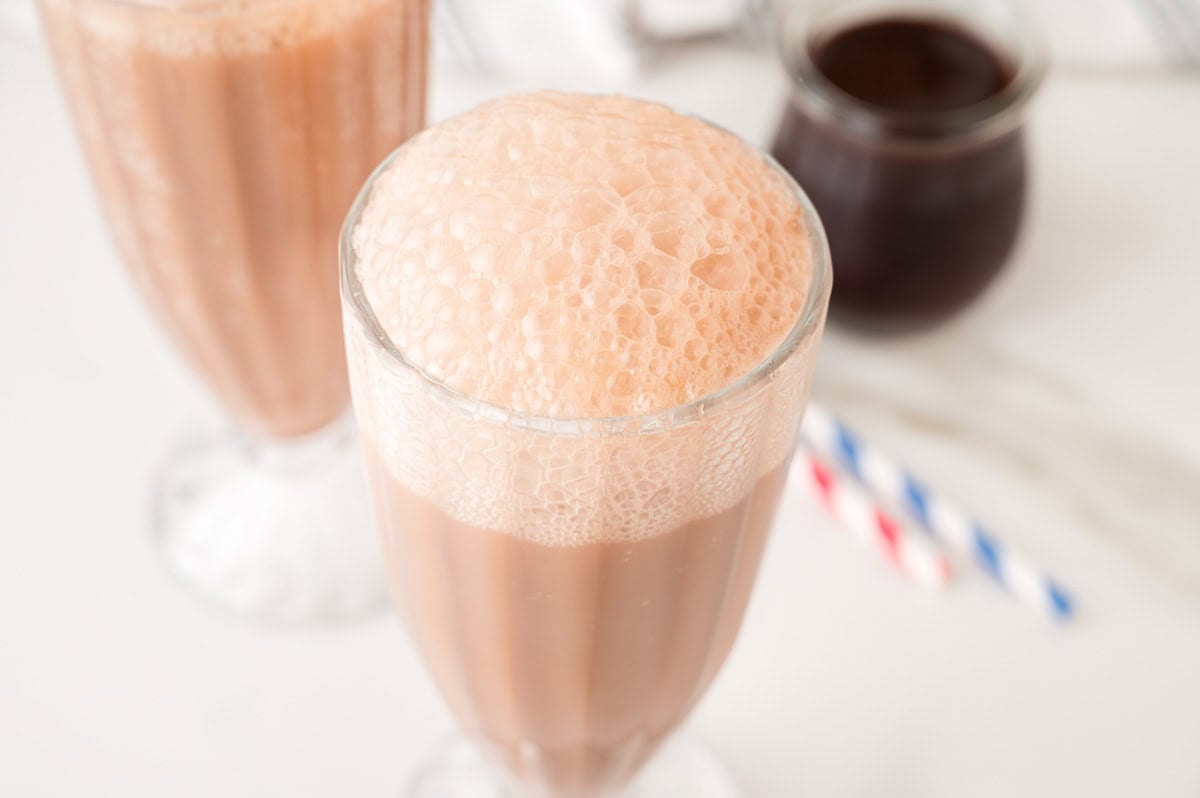 A glass of chocolate Egg Cream Soda with foam on top, with another glass and straws in the background.