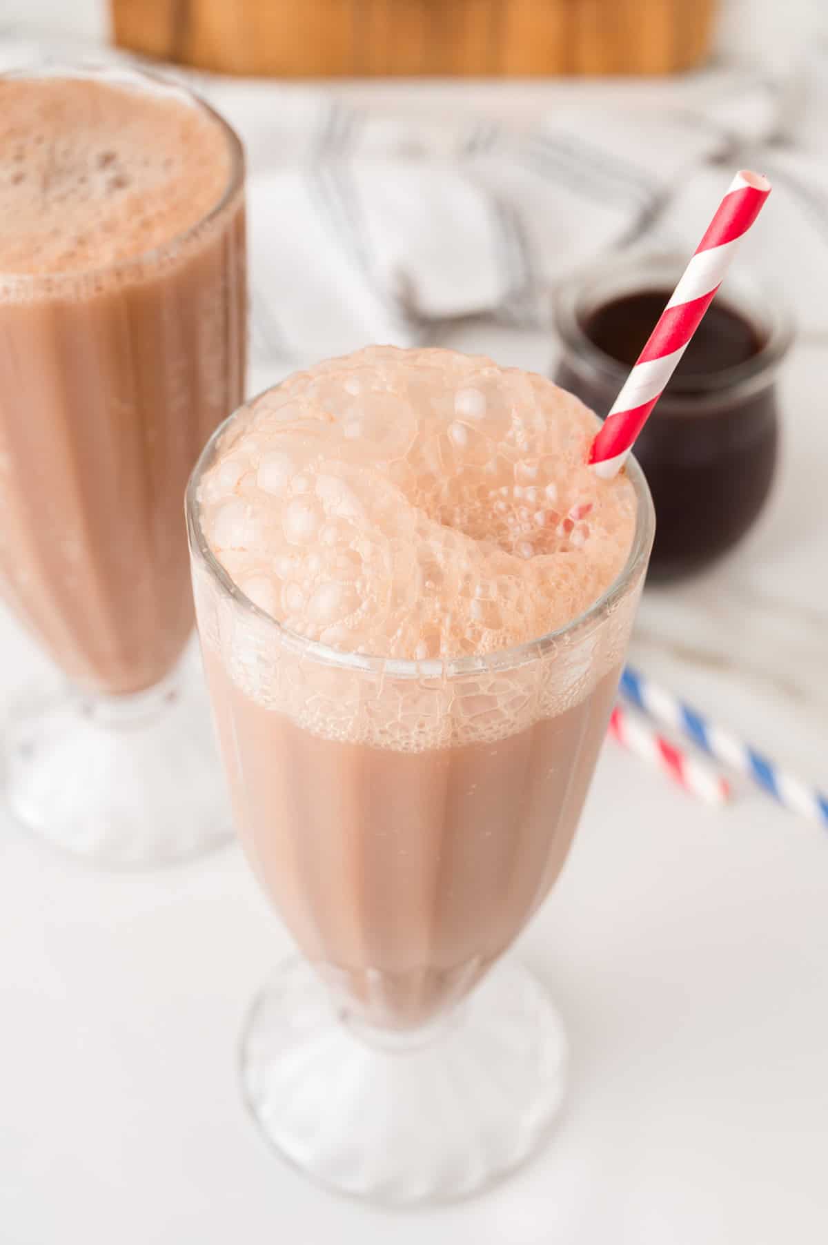 A frothy glass of chocolate milk with a red striped straw, reminiscent of a classic Egg Cream Soda, sits next to another glass and a jar in the background.