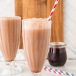 Two glasses of chocolate milk with red-striped straws, a jar of syrup, and a wooden board in the background evoke the classic charm of an old-fashioned Egg Cream Soda.