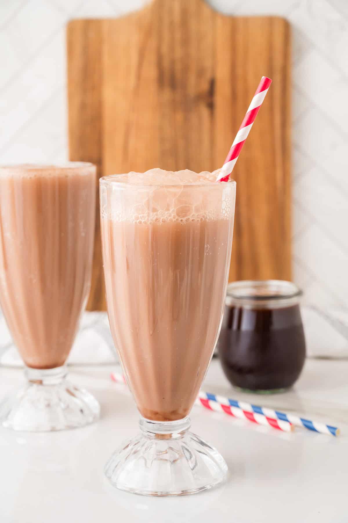 Two glasses of chocolate milk with red-striped straws, a jar of syrup, and a wooden board in the background evoke the classic charm of an old-fashioned Egg Cream Soda.