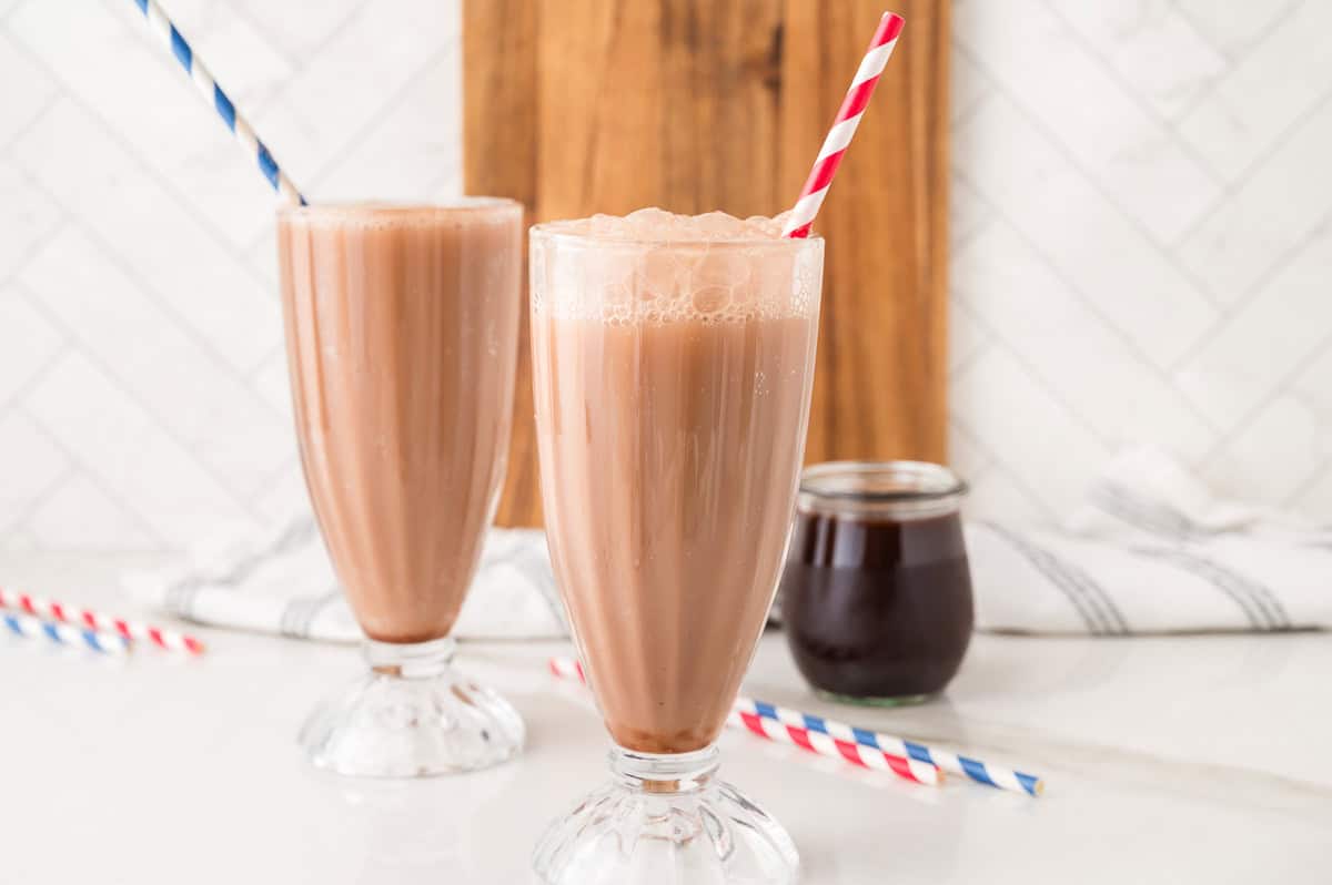Two chocolate milkshakes in tall glasses with straws, next to a jar of chocolate syrup on a white counter—an inviting setup reminiscent of an old-fashioned Egg Cream Soda shop.