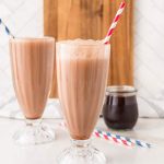 Two tall glasses of chocolate milk with red-striped straws sit on a white counter, reminiscent of classic Egg Cream Soda drinks, with a jar of chocolate syrup in the background.