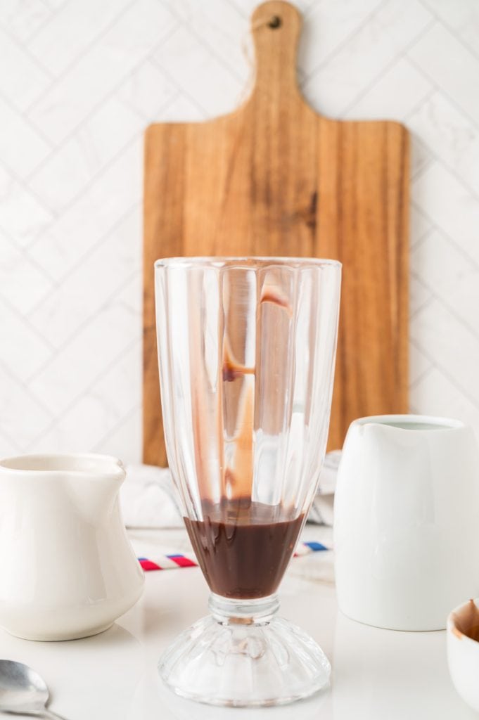 Empty glass with chocolate syrup inside, perfect for creating a classic Egg Cream Soda, set in front of a wooden cutting board and white pitchers.