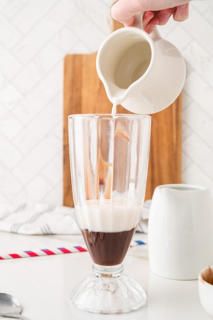 A hand pours milk from a small white pitcher into a tall glass with coffee on a kitchen counter, evoking the creamy swirl of an Egg Cream Soda.