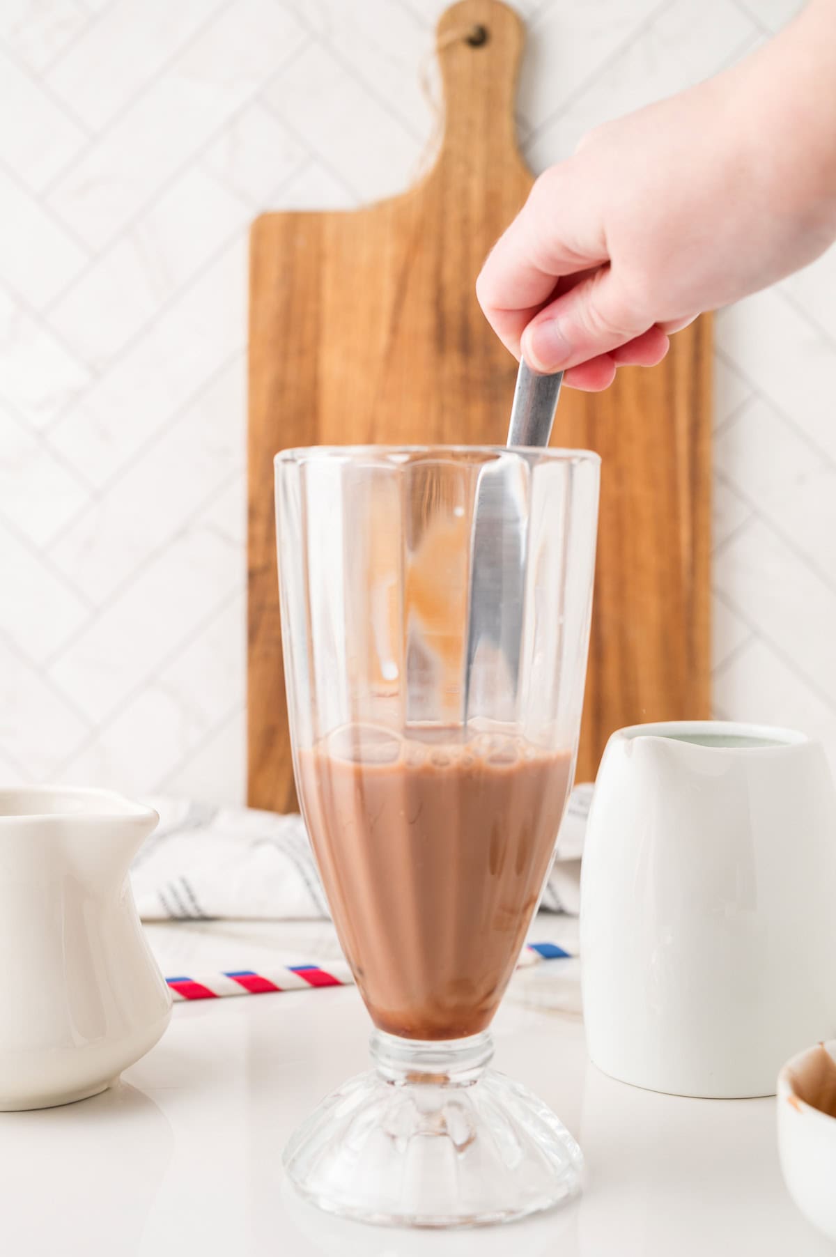 Hand stirring chocolate milk in a tall glass, reminiscent of crafting a classic Egg Cream Soda, with pitchers and a wooden board in the background.